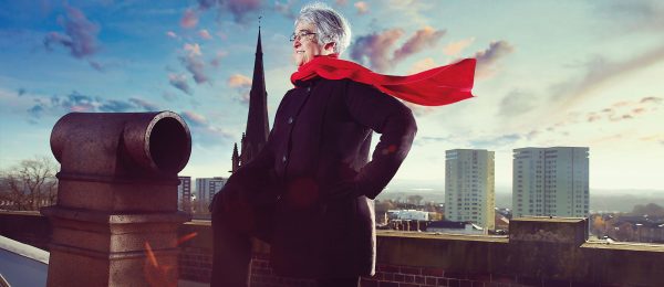 Female volunteer in her 60s on a rooftop, in superhero pose with red scarf blowing in the wind