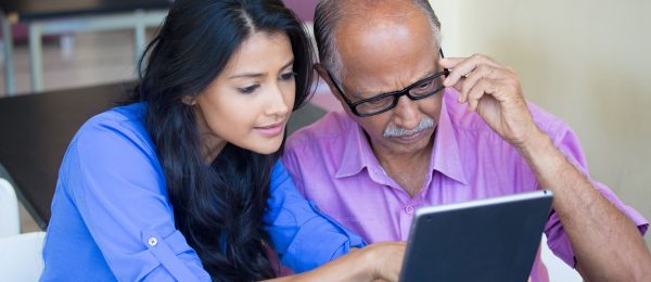 young woman sits with senior man helping him with tablet device